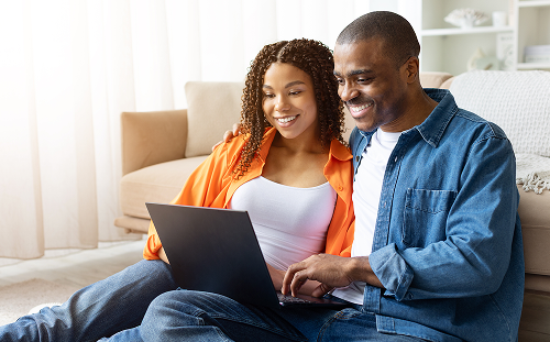 A smiling couple sits on the floor, using a laptop. They appear relaxed and happy in a cozy, bright room with soft furnishings in the background.
