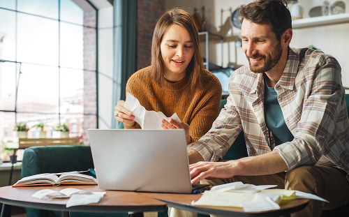 A smiling couple in casual clothes sits at a table with a laptop and papers, appearing relaxed and focused while managing finances in a bright living room.