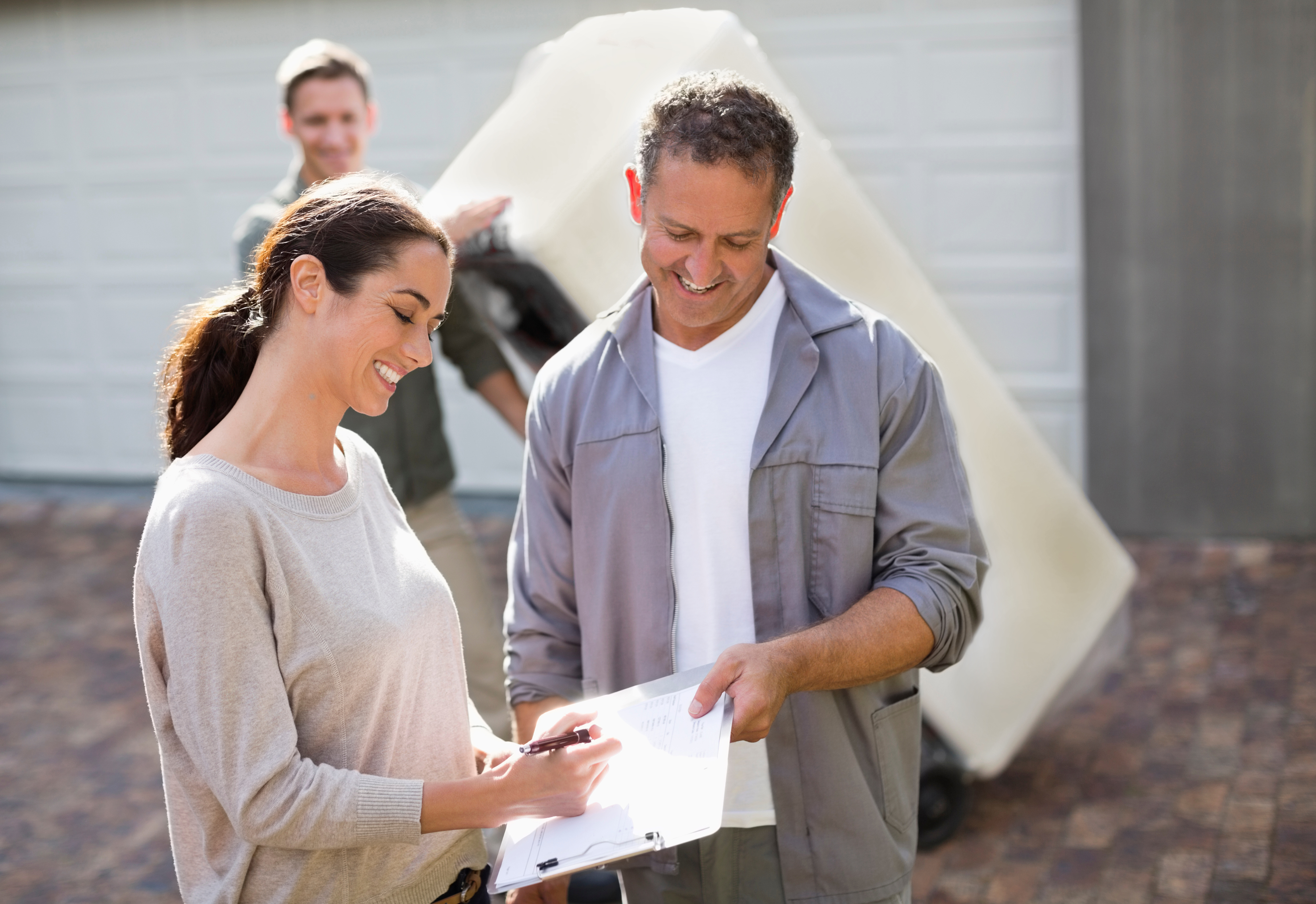 A smiling woman signs a document held by a man in workwear outside, while another man moves an appliance.