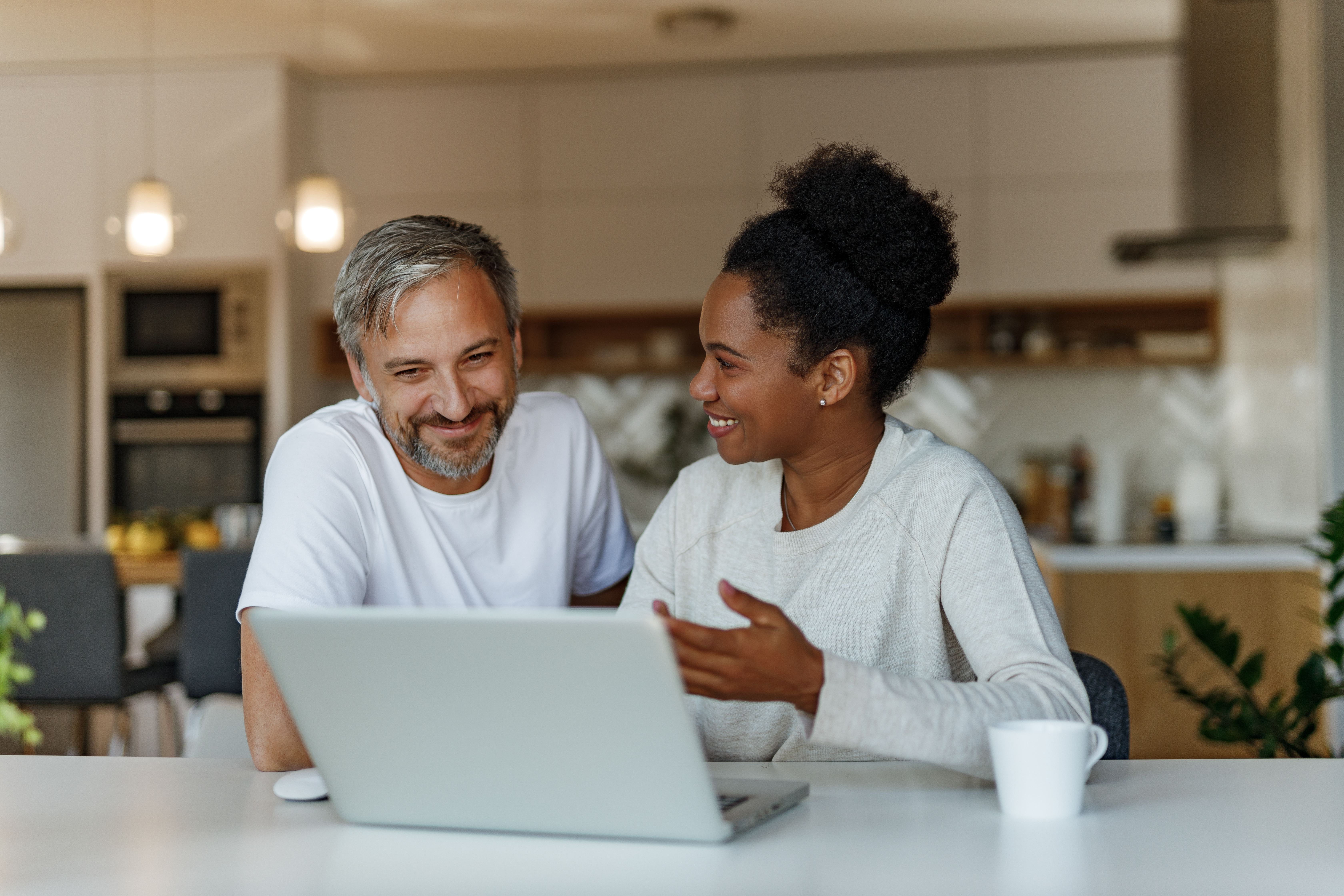 Two people sitting at a kitchen table using a laptop together in a home setting.