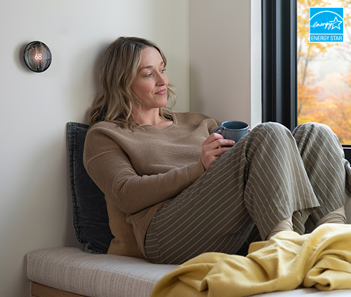 Woman relaxing on a window bench with a cup of coffee near a smart thermostat set to 68 degrees, with an ENERGY STAR logo in the corner.